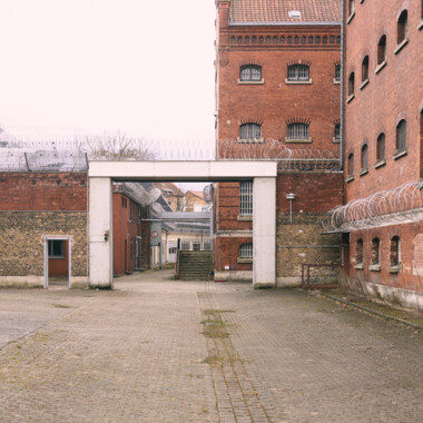 The paved inner courtyard of Rennelberg Prison. Between the brick buildings stands a wall with a large gate leading into one of the inner courtyards. There is barbed wire along the walls, with further buildings visible in the background.